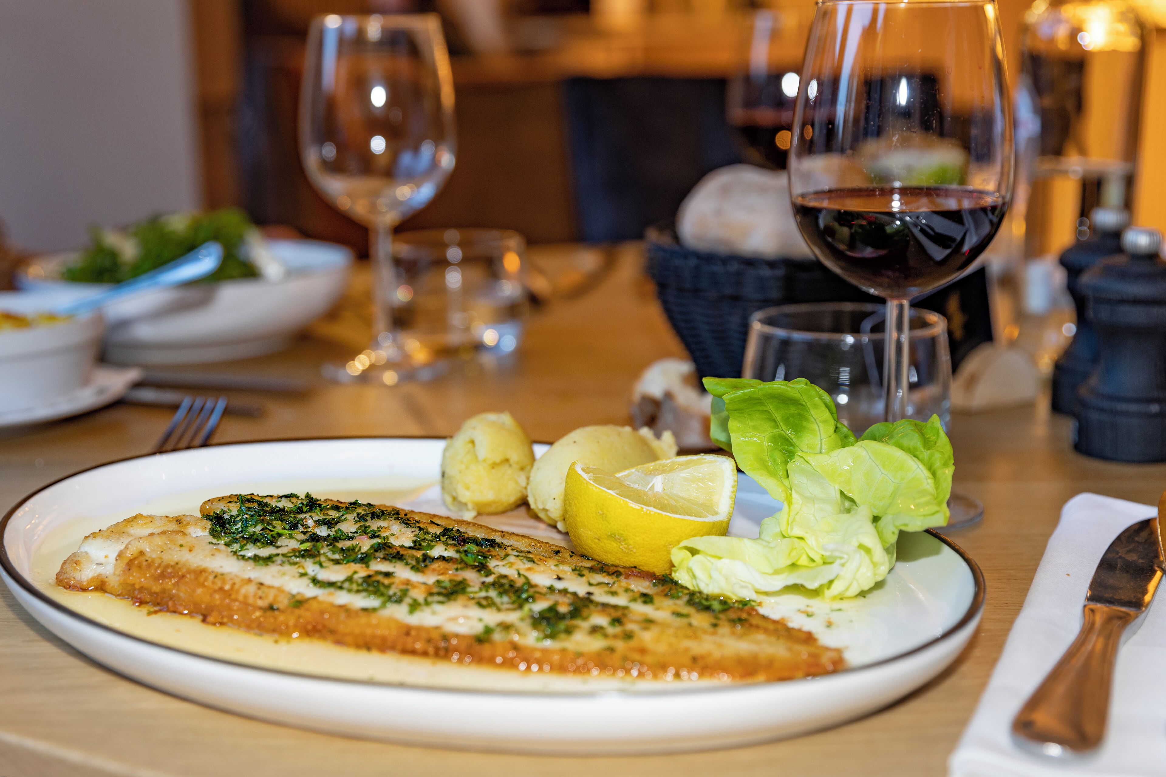 A plate with a fish fillet, lemon, and greens, and a glass of red wine on a table.