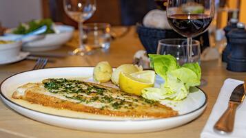 A plate with a fish fillet, lemon, and greens, and a glass of red wine on a table.