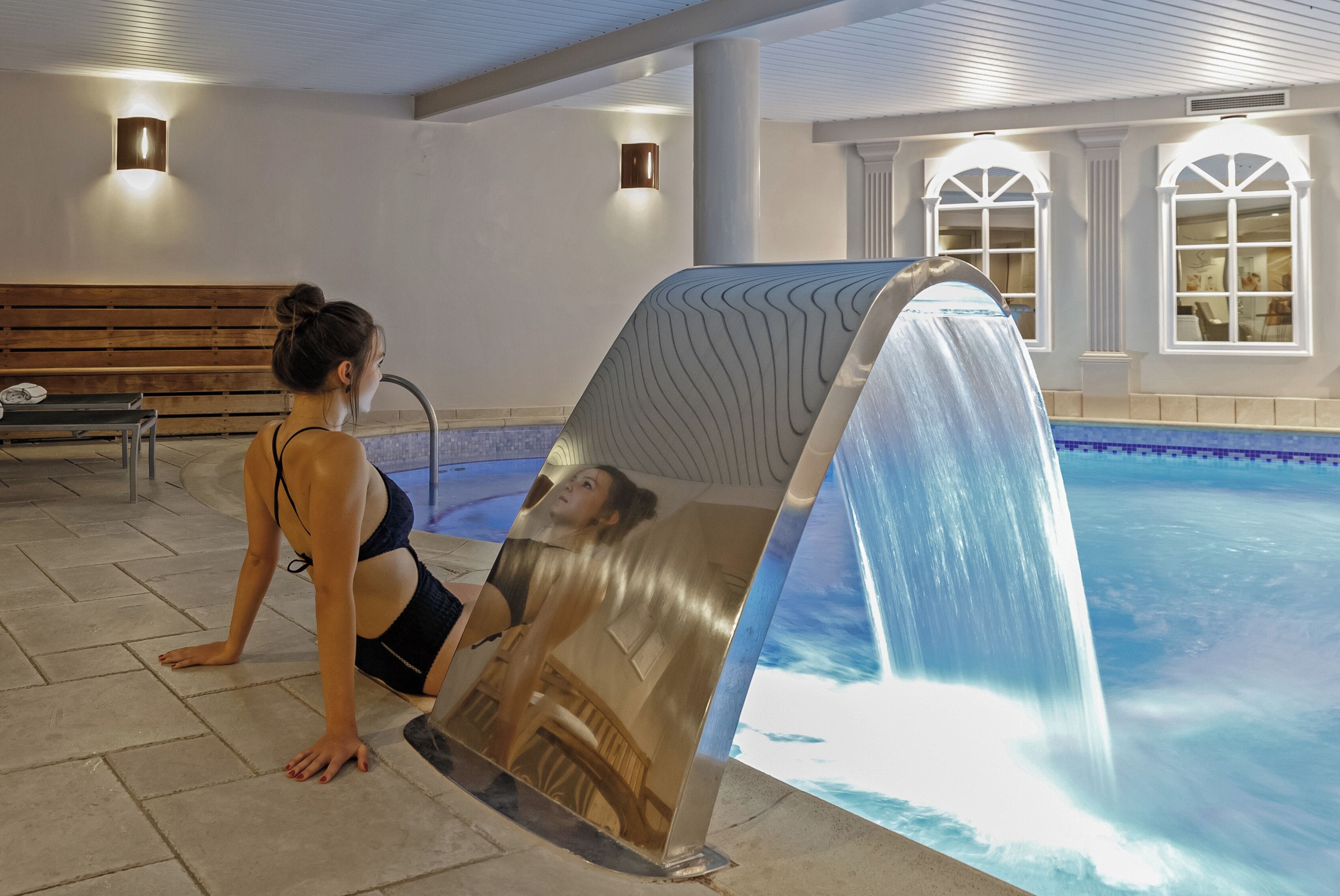 A woman in a swimsuit sitting by a pool with a waterfall feature.