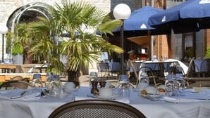 An outdoor dining area with tables set for two, covered by umbrellas, and a palm tree in the center. - Hotel Quartier Latin (Marche-en-Famenne)