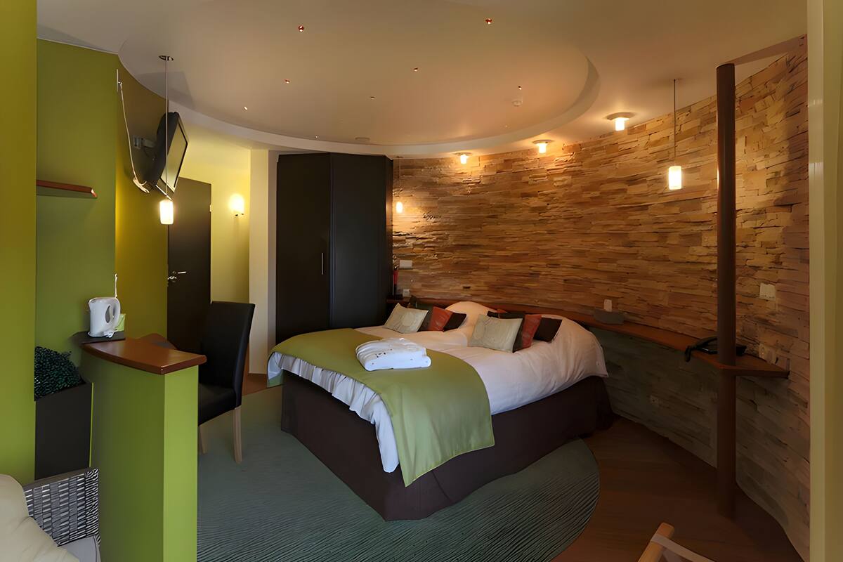 A modern bedroom with a curved ceiling, a stone accent wall, a bed with a green throw, and a bedside table with a kettle.