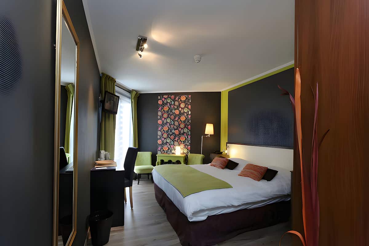 A modern hotel room with a large bed, a desk with a computer, a chair, and a small table with a lamp.