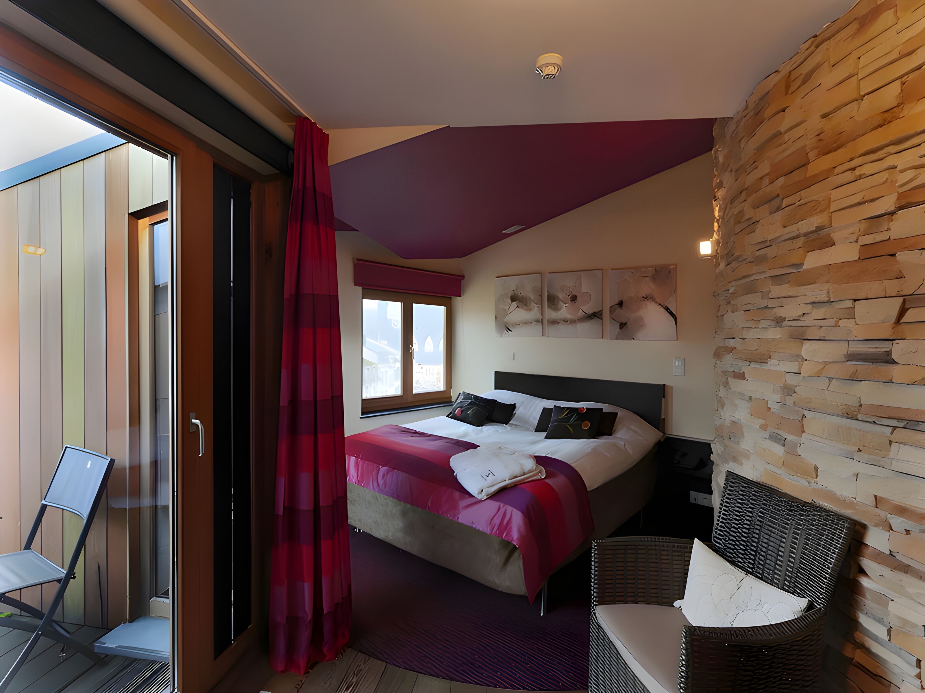 Une chambre à coucher comprenant un lit, une chaise, une fenêtre et un mur à la texture rappelant celle de la pierre.