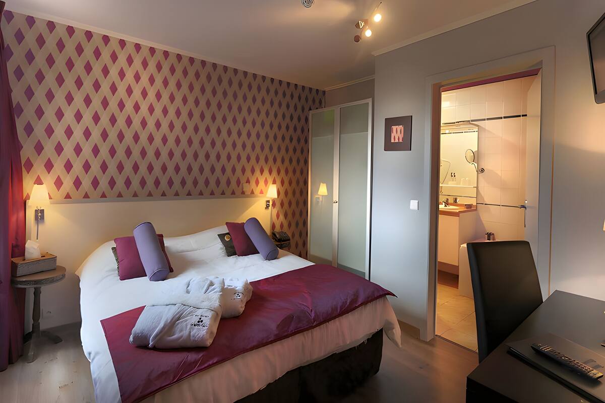 A hotel room with a large bed, a desk with a chair, a bathroom visible through an open door, and a patterned wall.