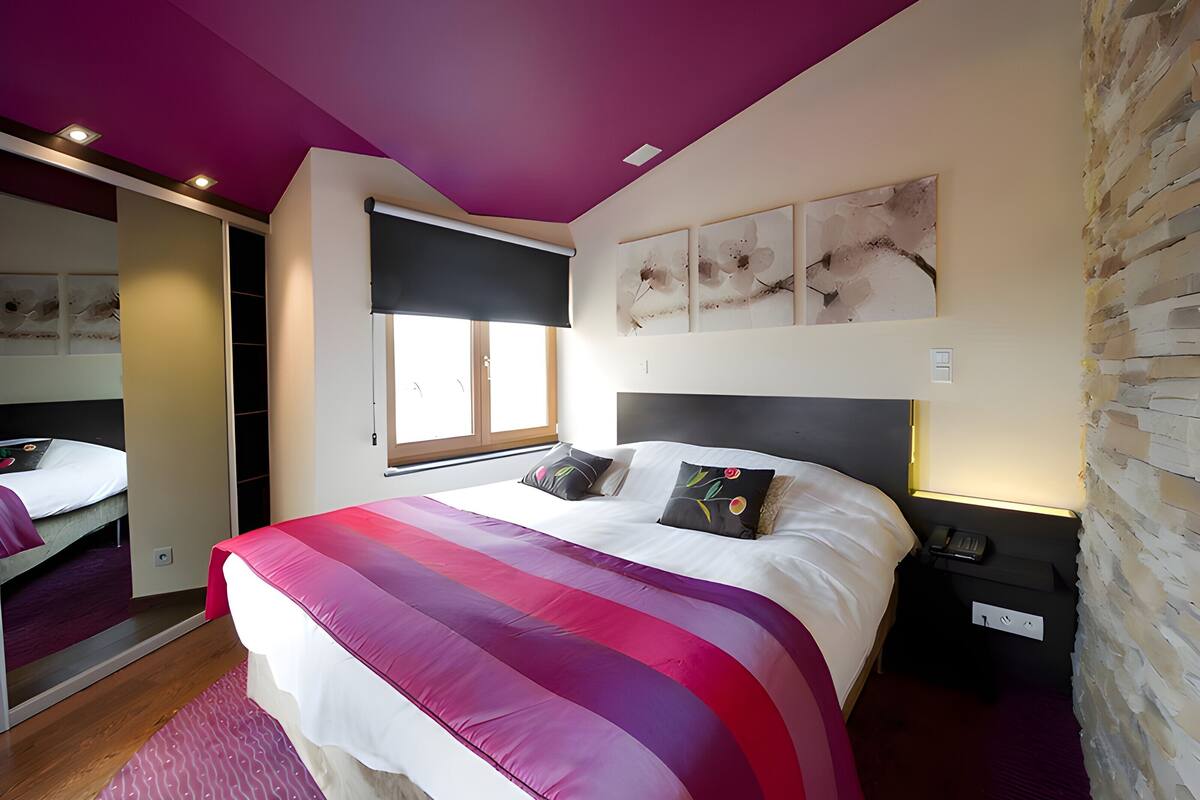 A modern bedroom with a large bed, a window with blinds, a stone accent wall, and a built-in wardrobe.