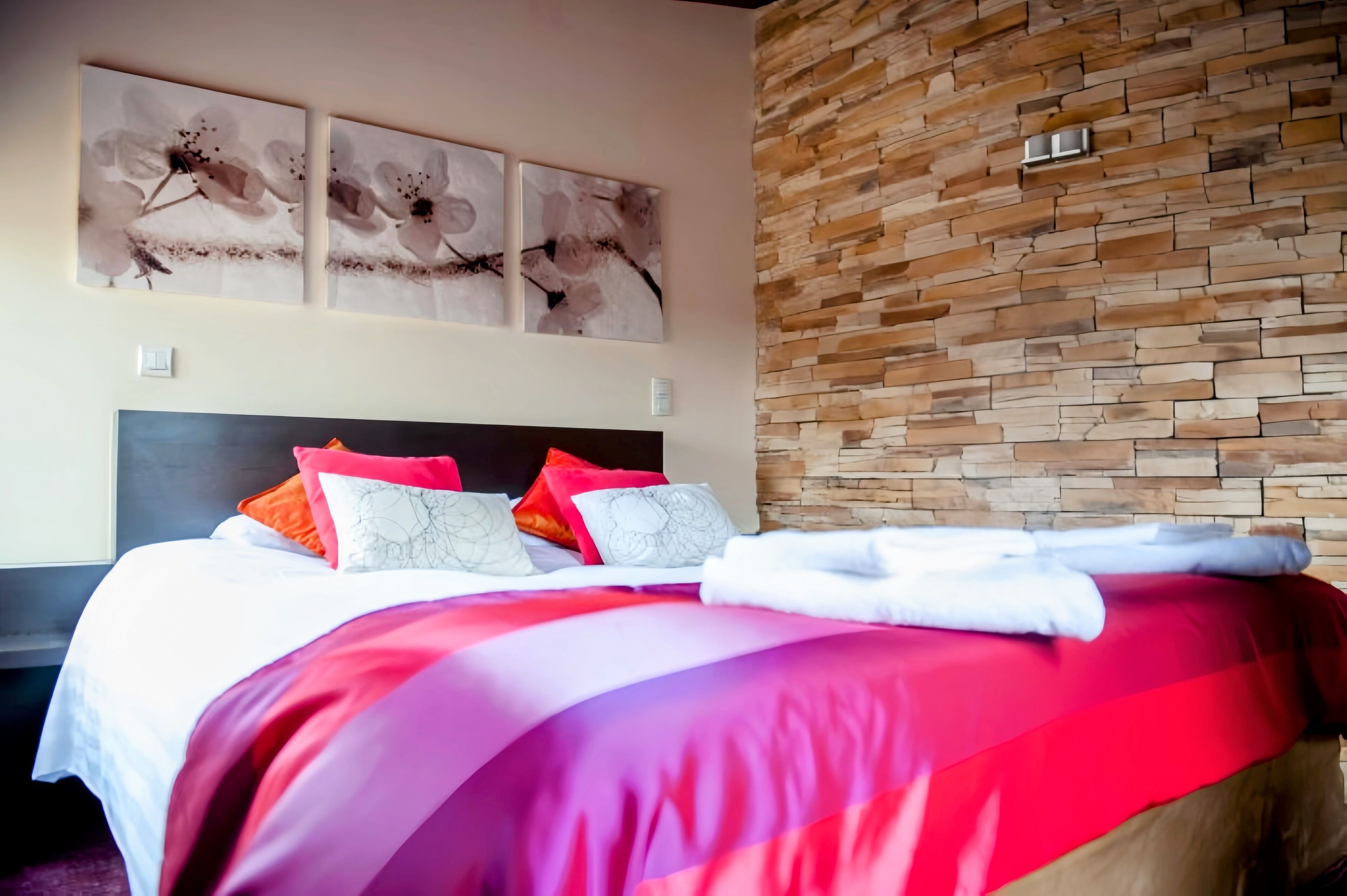 Una habitación de hotel con pared de piedra, una cama con ropa de cama roja y blanca, y tres cuadros florales en la pared.