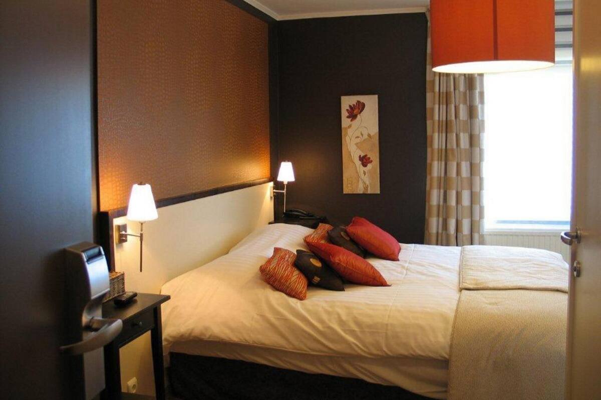 A hotel room with a bed, two bedside lamps, a desk with a computer, and a window with curtains.