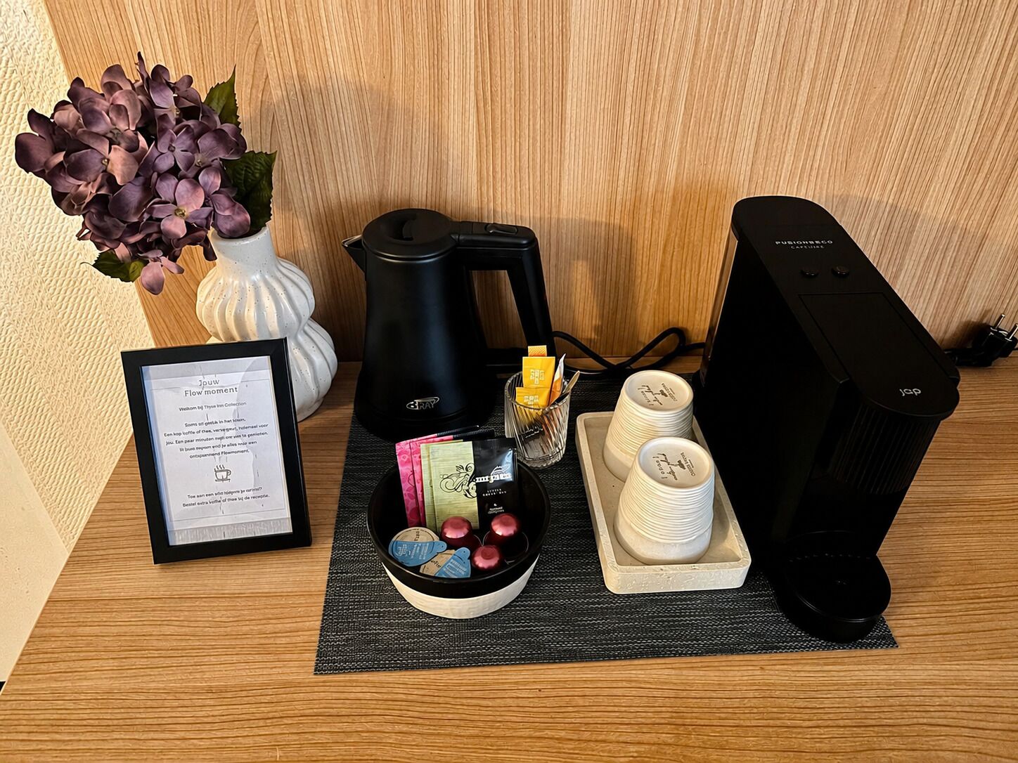 Een opgeruimd werkplek met een koffiezetapparaat, een computer, een ingelijst diploma, een vaas met bloemen en een dienblad met koffiekopjes en een mok.