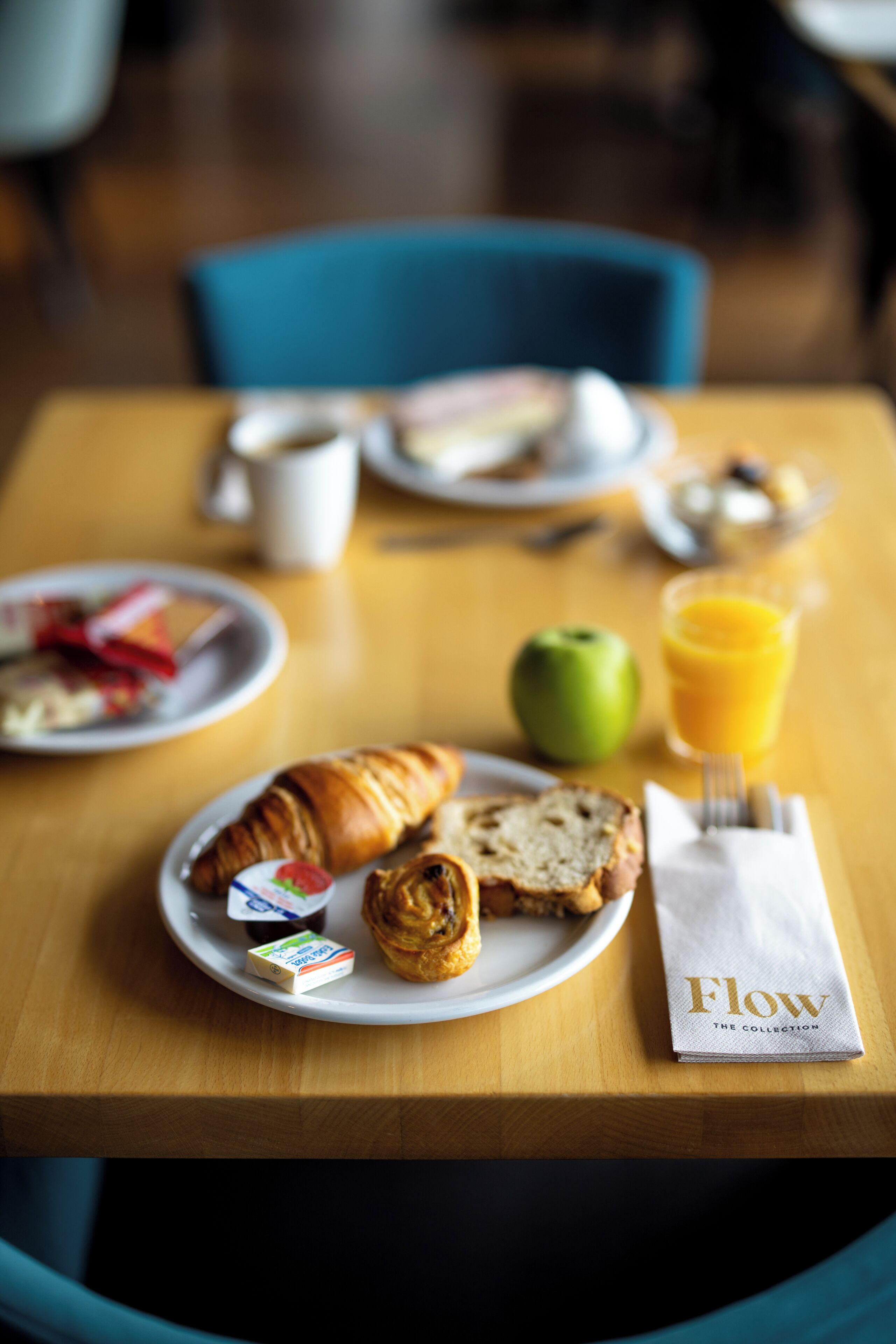 Ein Frühstückstisch mit einem Croissant, einem Muffin, einem Glas Orangensaft, einem Teller mit einem grünen Apfel und einer Serviette mit dem Aufdruck 'Flow'.