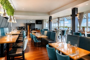 A restaurant with wooden tables and chairs, a clock on the wall, and large windows offering a view of the water. - Hotel Hoogeerd by Flow (Niftrik)