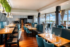 Um restaurante com mesas e cadeiras de madeira, um relógio na parede e grandes janelas com vista para a água.