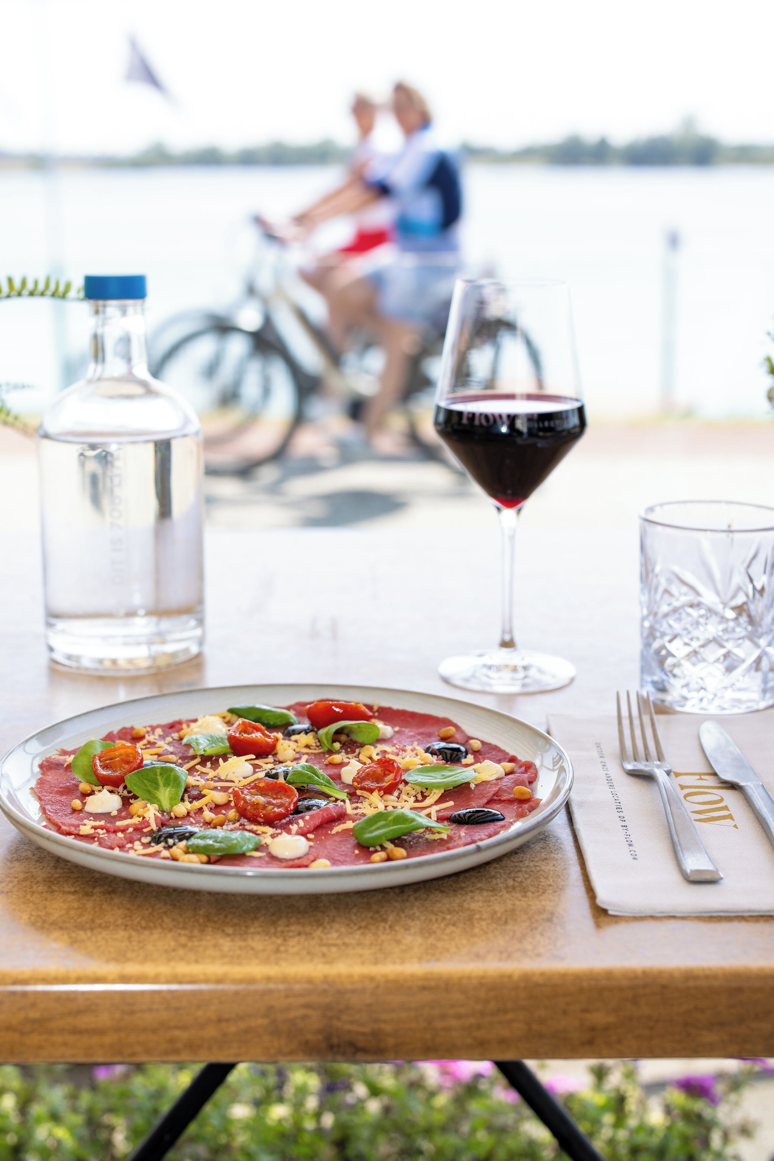 Una mesa de comedor con un plato de comida, una copa de vino tinto, una botella de agua y un vaso de agua.