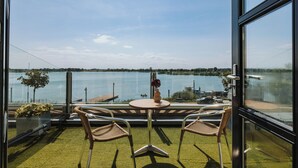 Een kamer met uitzicht op een meer, twee rieten stoelen, een klein rond tafeltje en een vaas met bloemen.