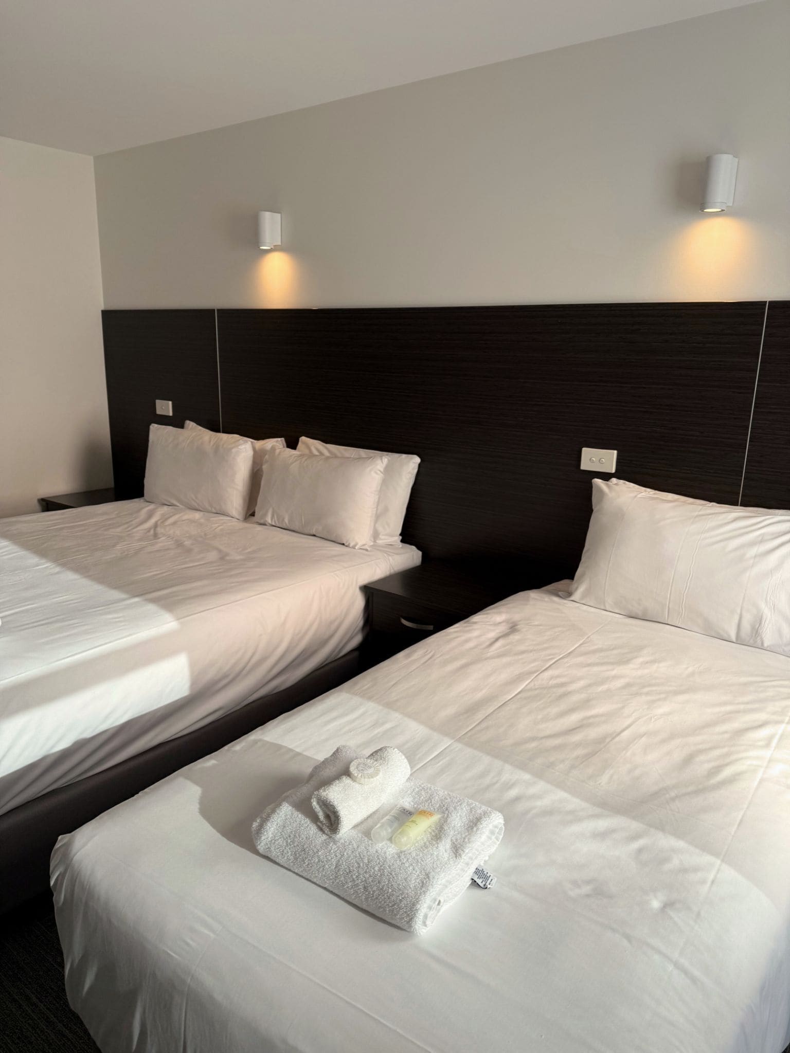 A hotel room with two beds, each with white linens and pillows. There are wall-mounted light fixtures above each bed. A small table with a towel and toiletries is visible in the foreground.