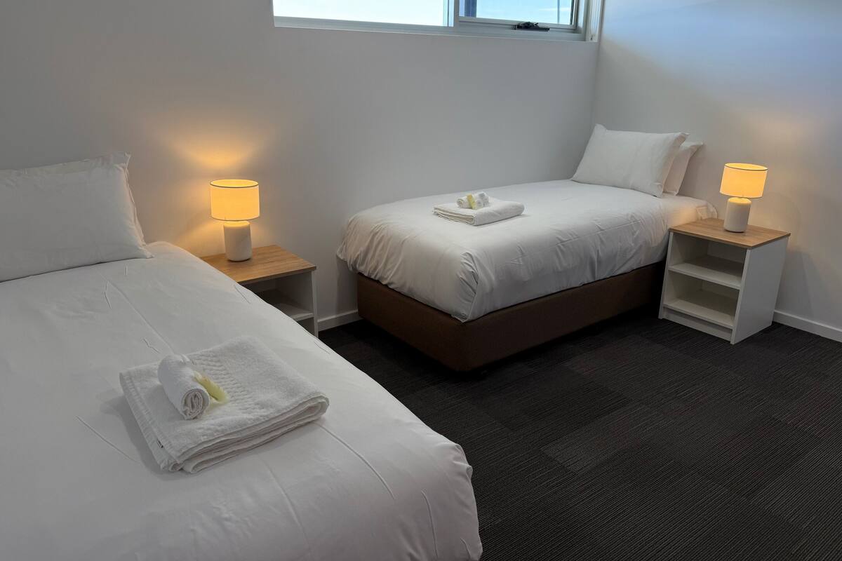 A hotel room with two beds, each with a bedside table and lamp, a window, and a carpeted floor.