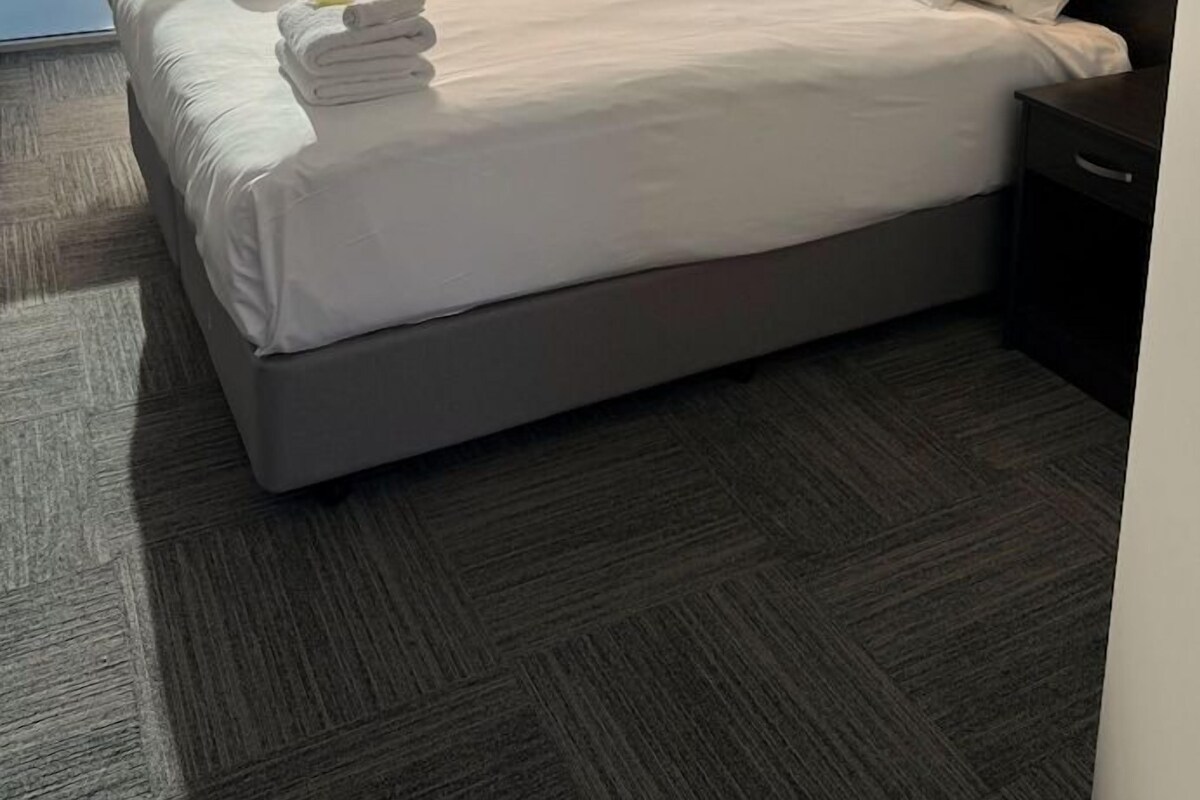 A hotel room with a large bed, a nightstand, a wall-mounted light, and a view through a sliding door.