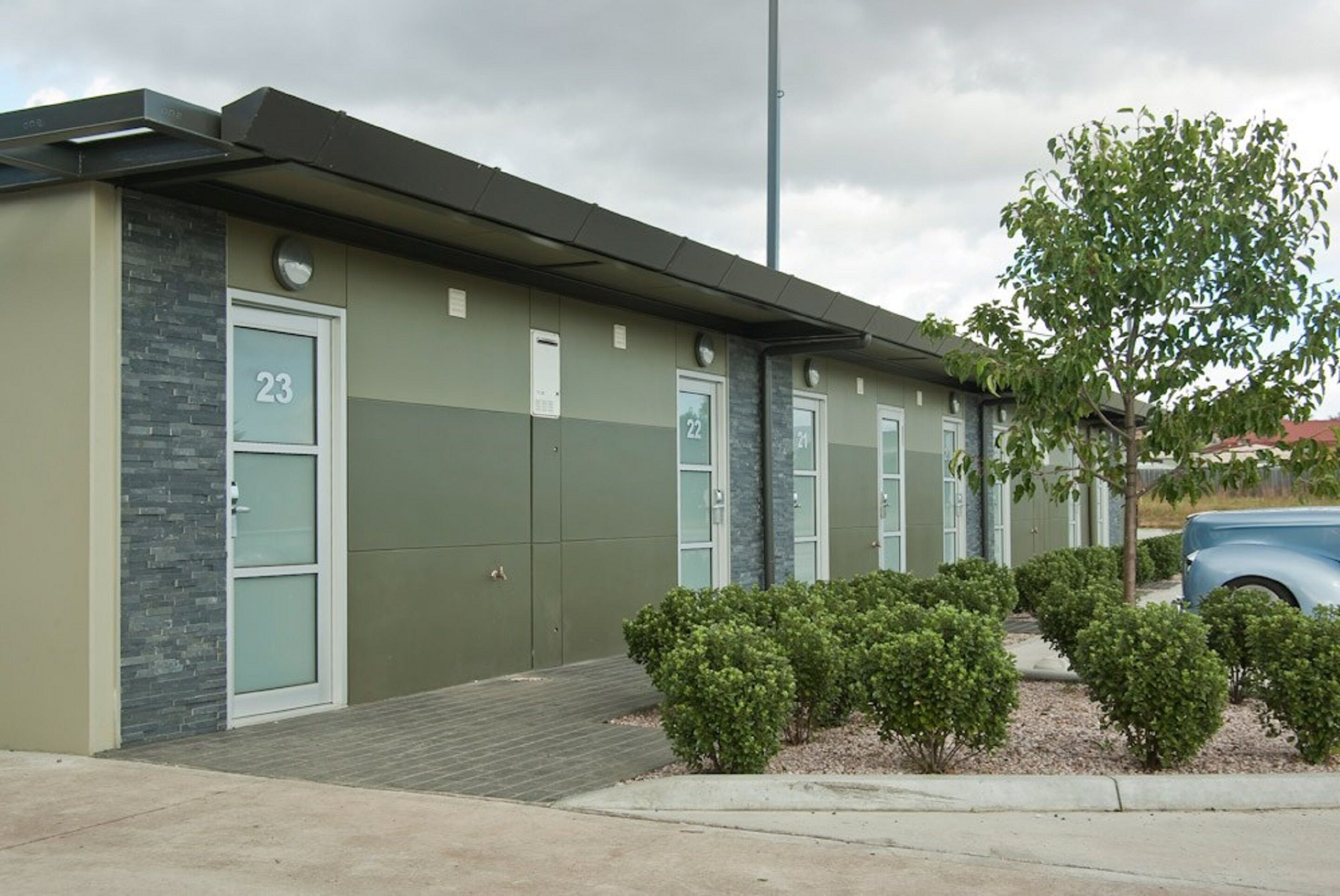 A building with numbered doors, a small entrance, and a parking area with a tree and bushes.