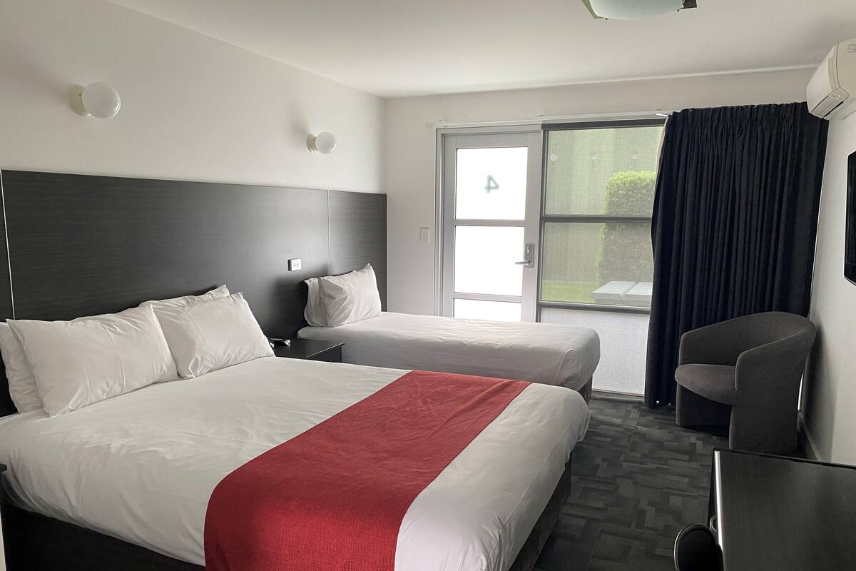 A hotel room with a large bed, a smaller bed, a desk, and a chair. There is a window with curtains and a view of the outdoors.