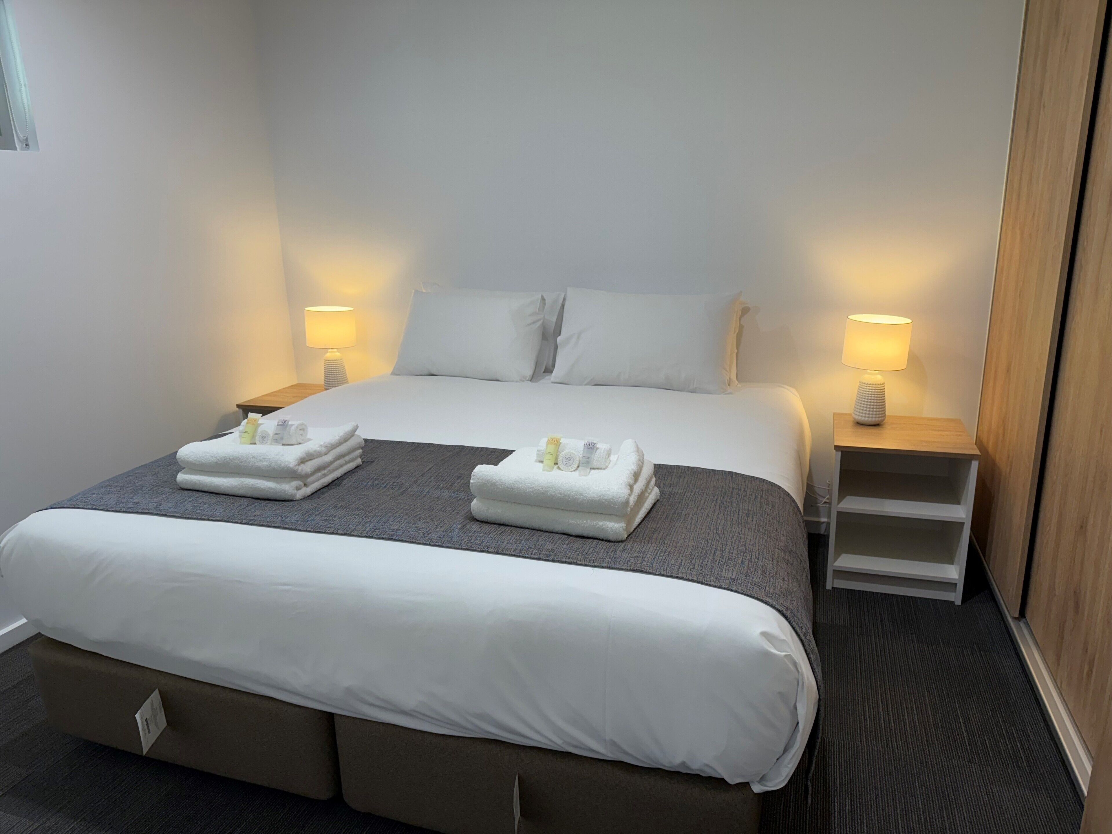 A hotel room with a large bed, two bedside tables with lamps, a small shelf, and a carpeted floor.