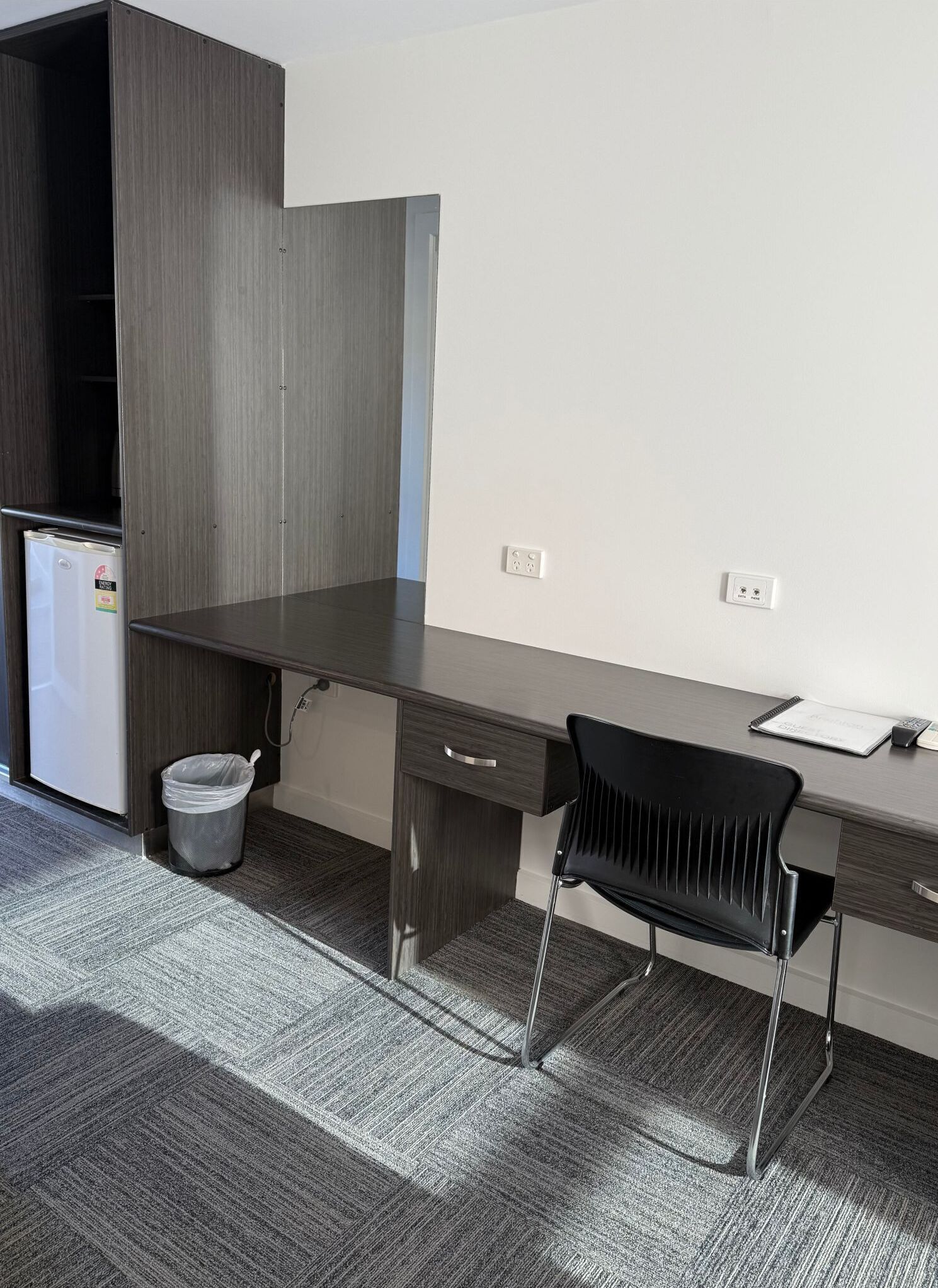 A compact office space with a dark wooden desk, a black chair, a white trash bin, and a small white refrigerator.