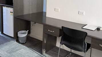 A compact office space with a dark wooden desk, a black chair, a white trash bin, and a small white refrigerator.