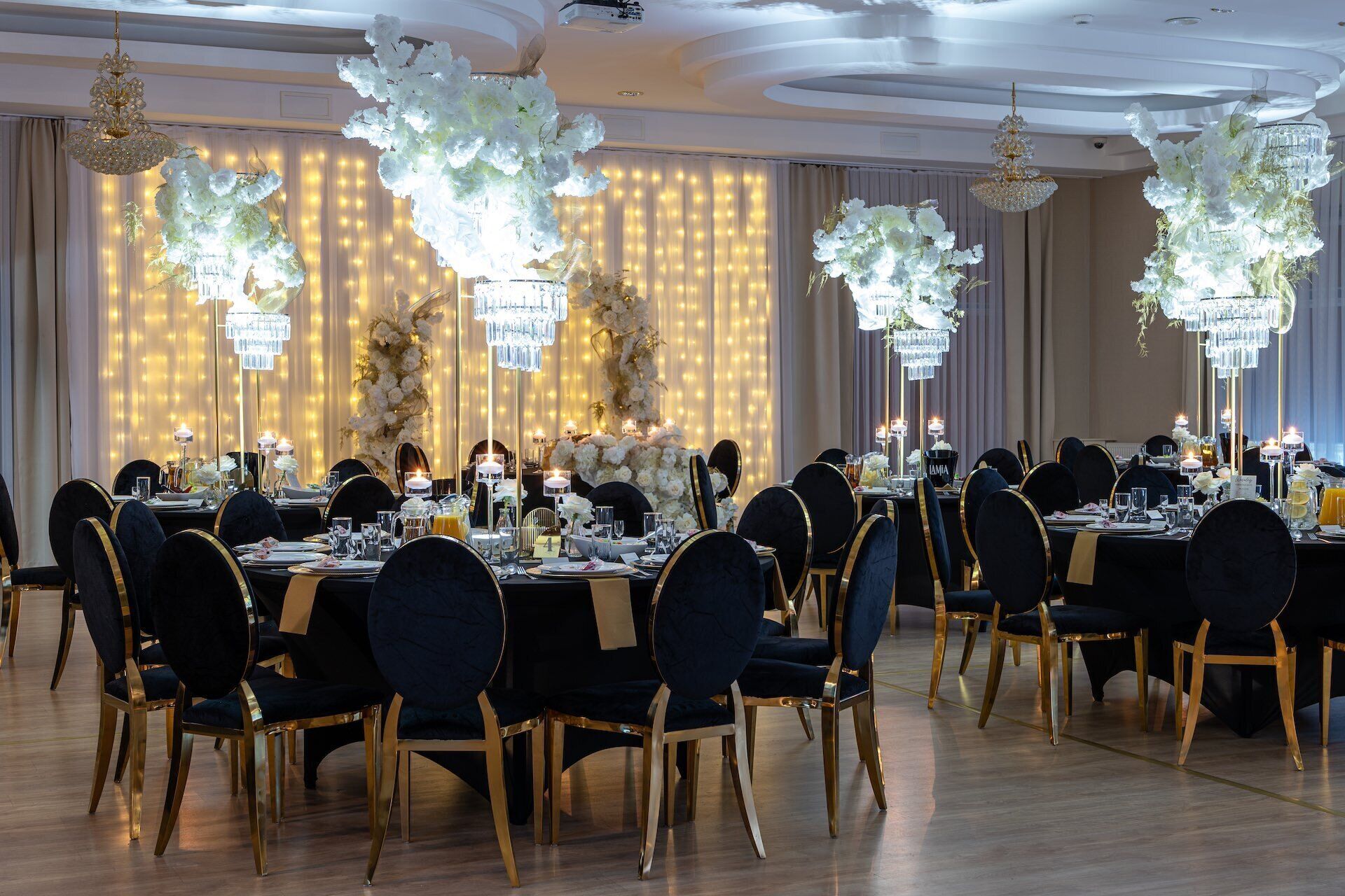 A formal dining room with tables set for multiple guests, chandeliers, and a backdrop of curtains.