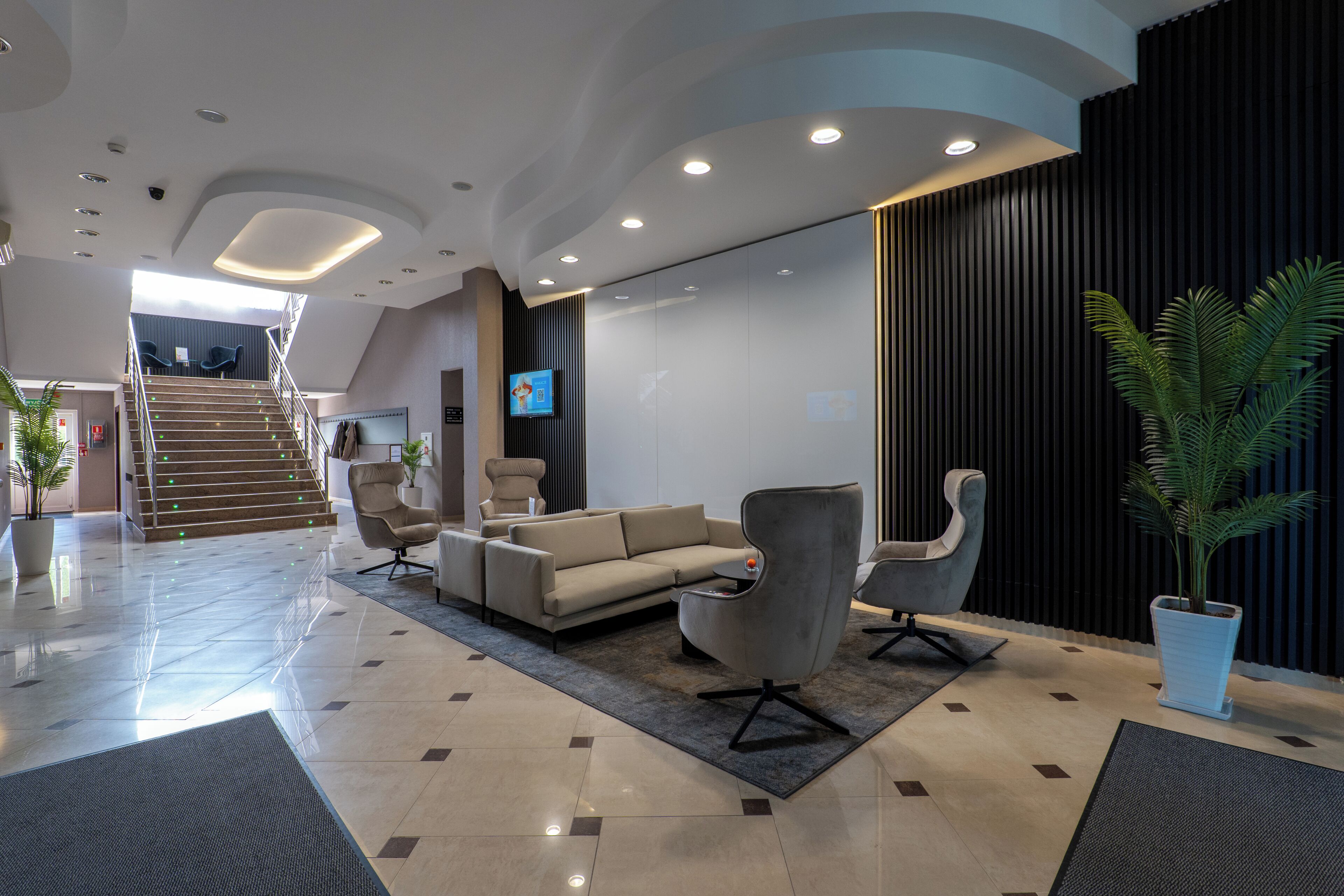 A modern lobby with a sofa, armchairs, a staircase, and a TV screen.