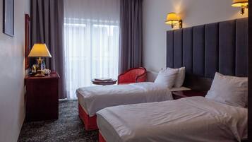 A hotel room with two beds, a red chair, a bedside table with a lamp, and a window with curtains.
