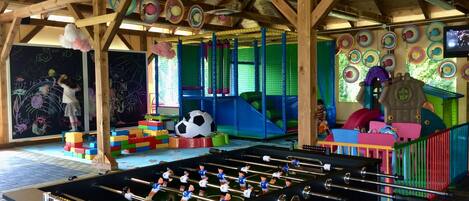 Zona infantil con mesa de fútbol de mesa, estructuras de juego coloridas y una pared de pizarra.