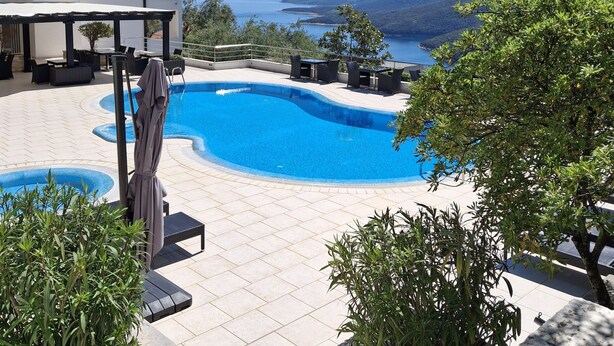 Piscina com vista para o mar e colinas, rodeada por áreas de estar ao ar livre e vegetação.