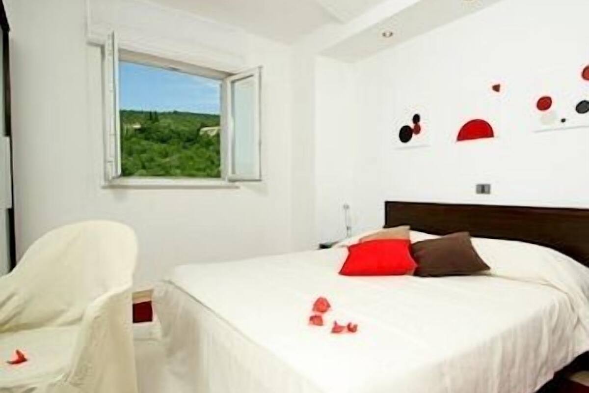 Une chambre à coucher avec un lit, une chaise, une fenêtre donnant sur un paysage verdoyant et des œuvres d’art mural.