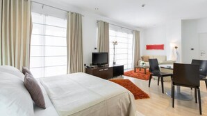Quarto de hotel moderno com uma cama grande, uma mesa com televisão e uma área de refeições com cadeiras e mesa.