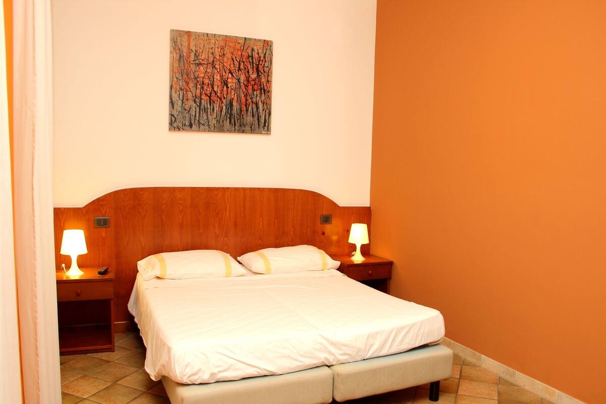 A hotel room with a wooden headboard, a bed with white bedding, two bedside lamps, and a painting on the wall.