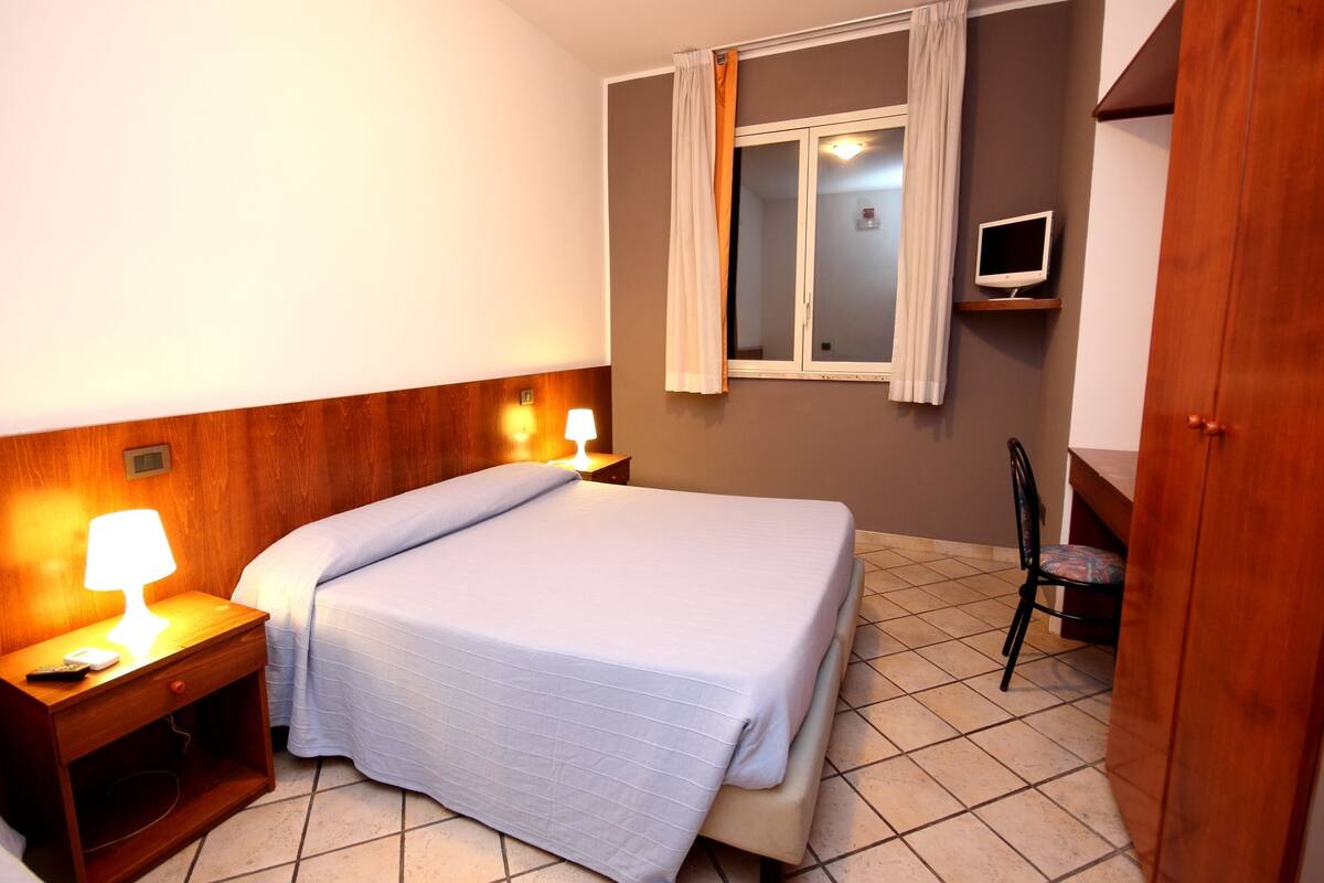 A hotel room with a bed, a nightstand with a lamp, a TV mounted on the wall, and a wooden wardrobe.
