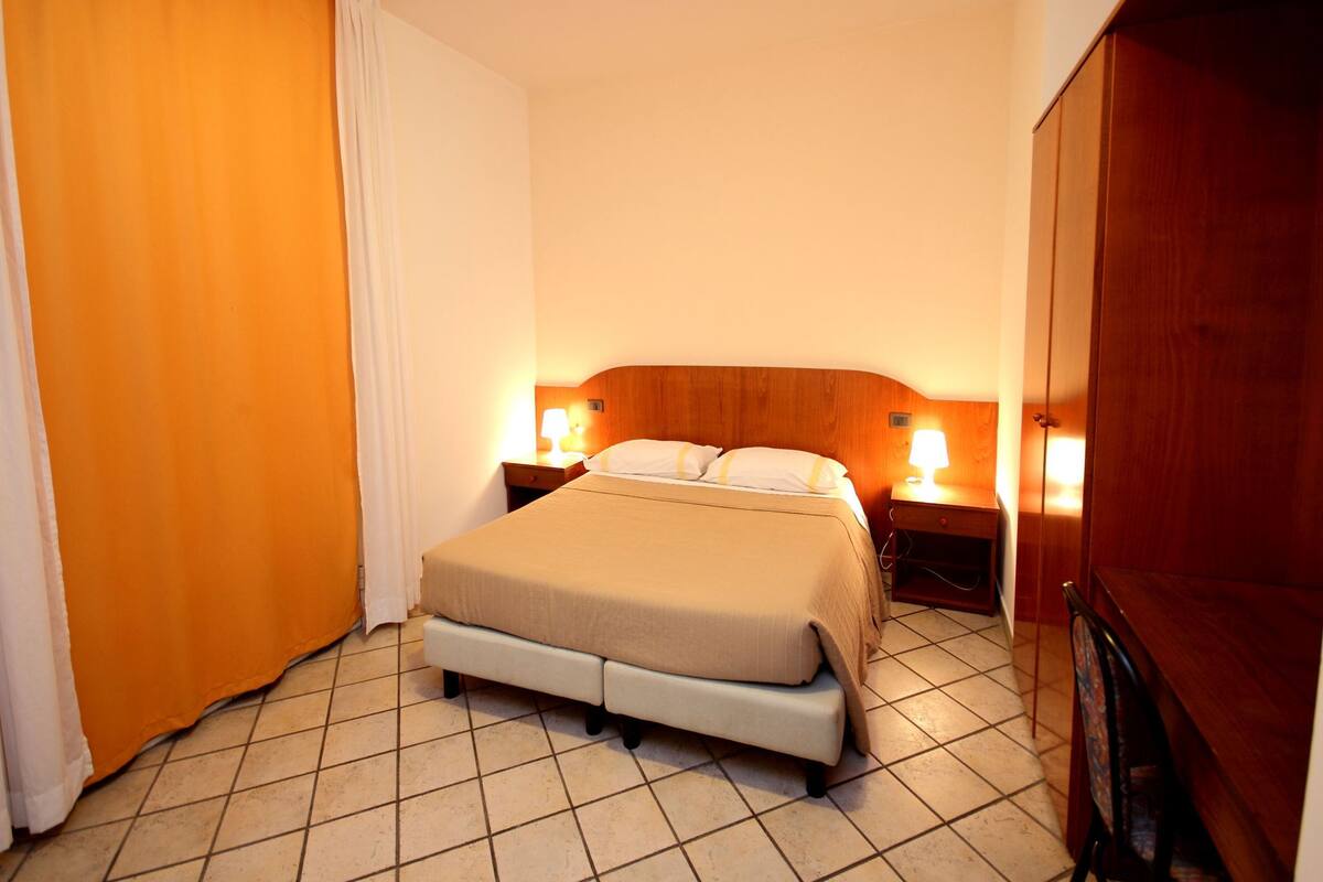 A hotel room with a large bed, two bedside tables with lamps, a wooden wardrobe, and a tiled floor.
