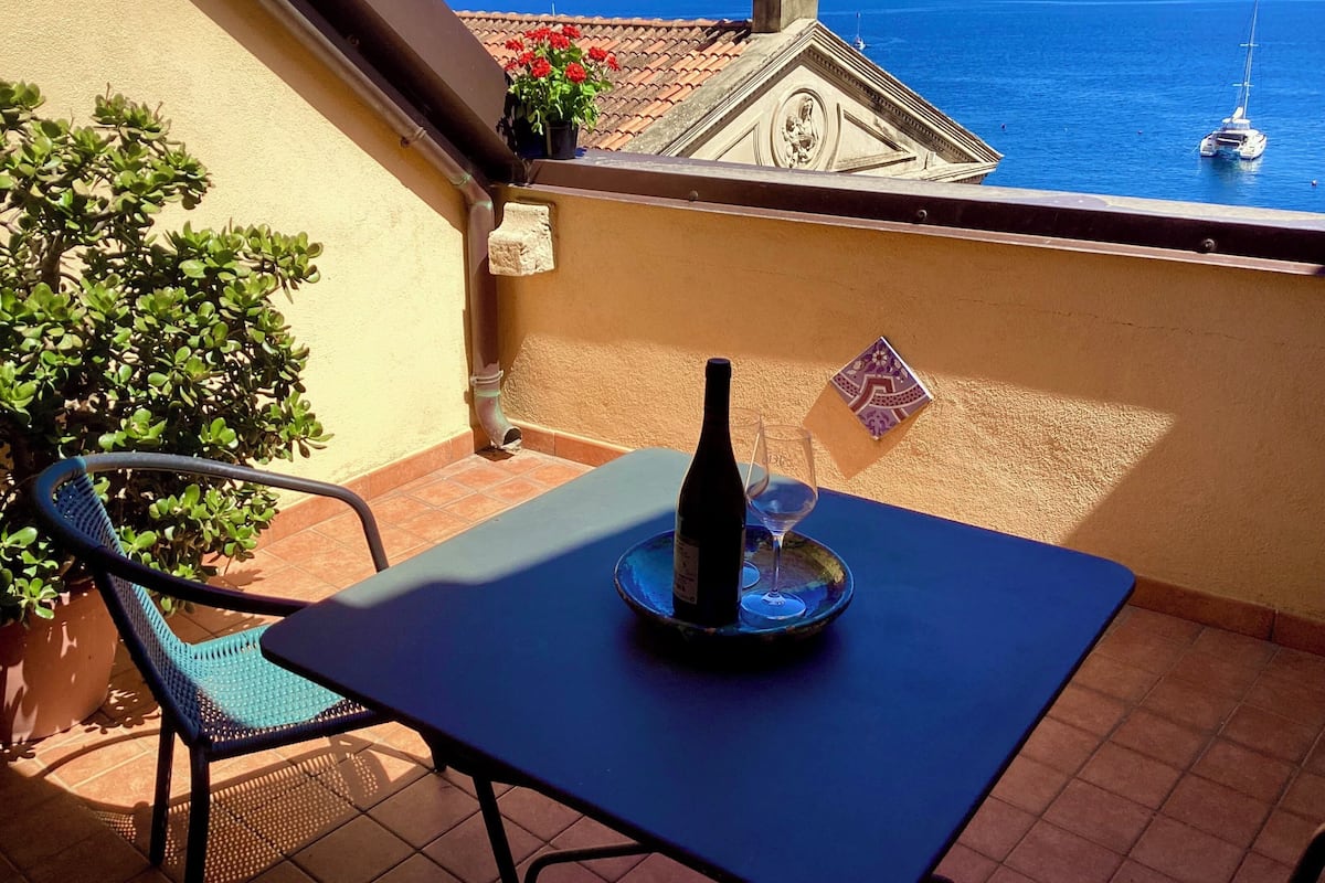 A balcony with a table, a chair, a bottle of wine, and a view of the sea.