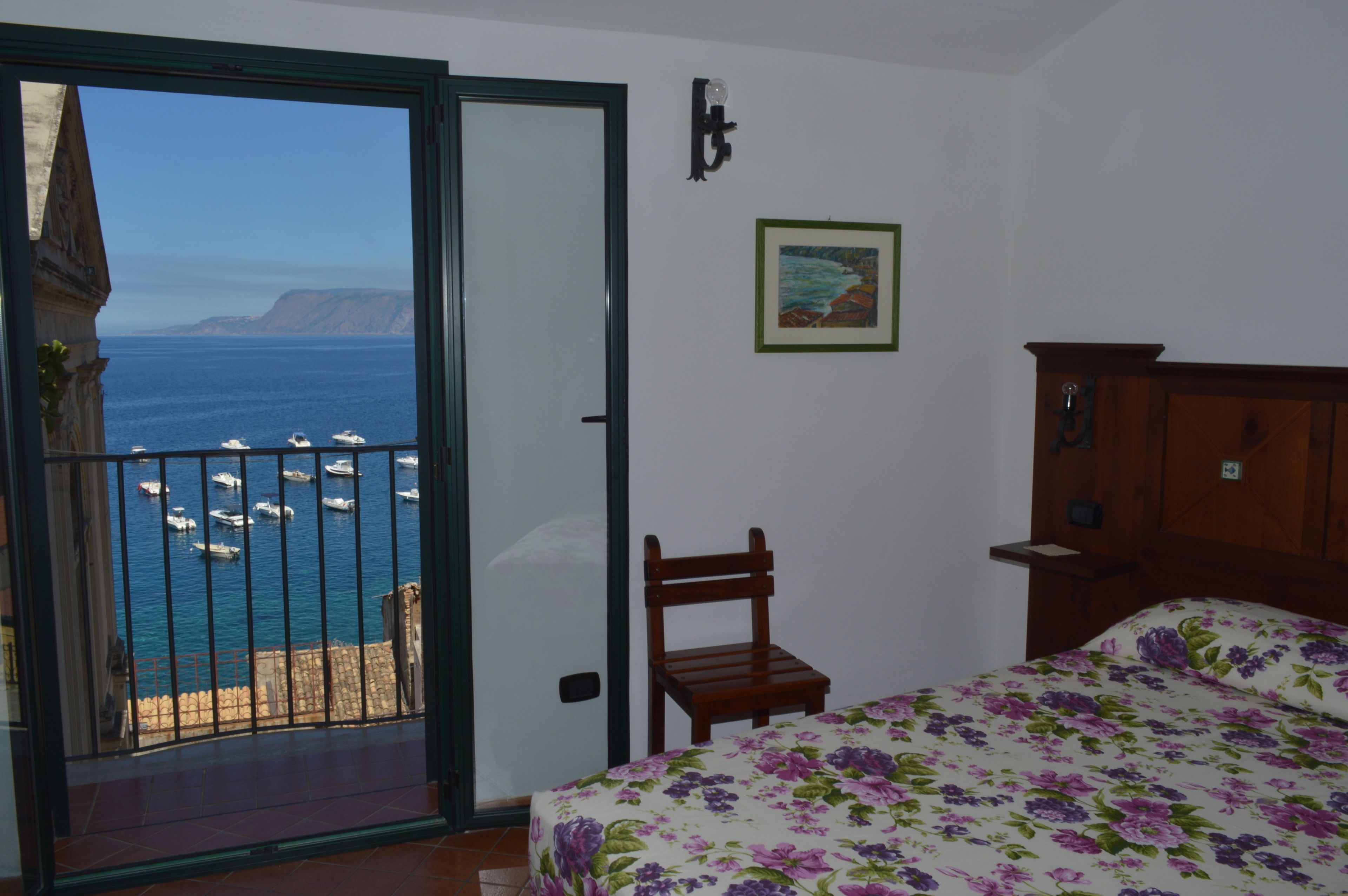 A bedroom with a bed, a chair, a wooden nightstand, a painting, and a view of the sea through an open door.