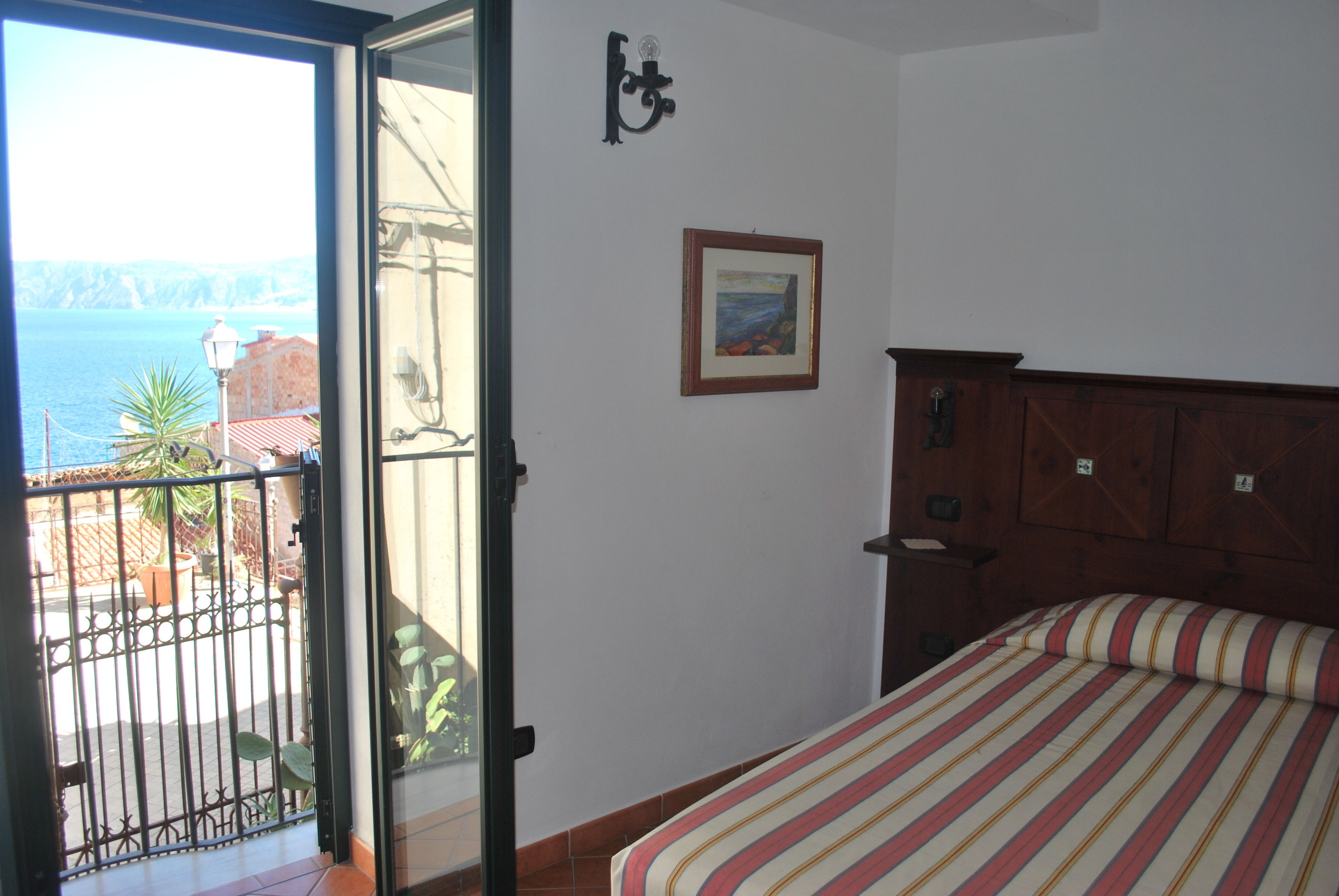Una camera da letto con un letto, un comodino, un armadio in legno, un quadro appeso al muro e un balcone con vista sul mare.