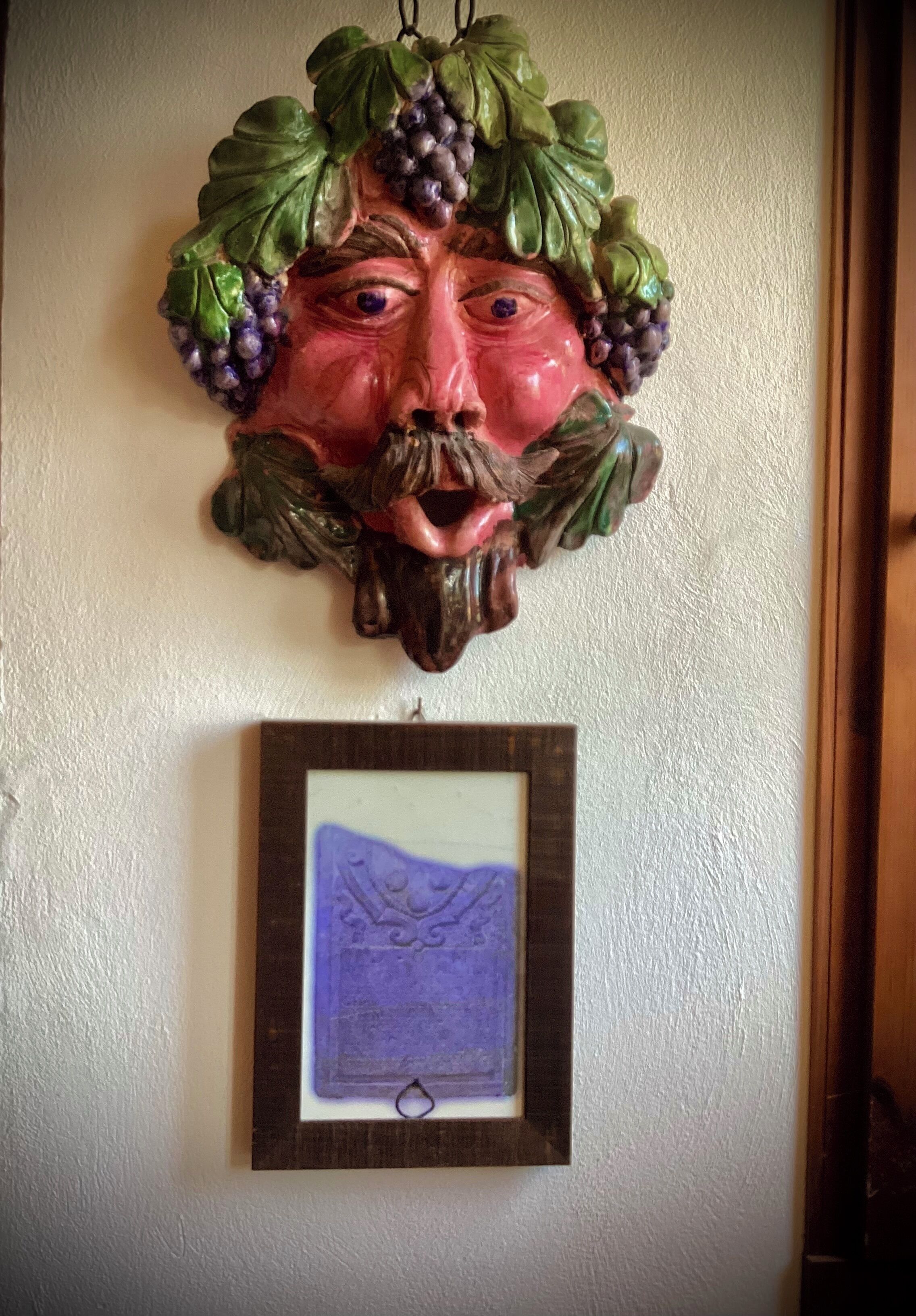 Un objet décoratif mural représentant un visage et des motifs de vigne, avec un tableau encadré placé en dessous.