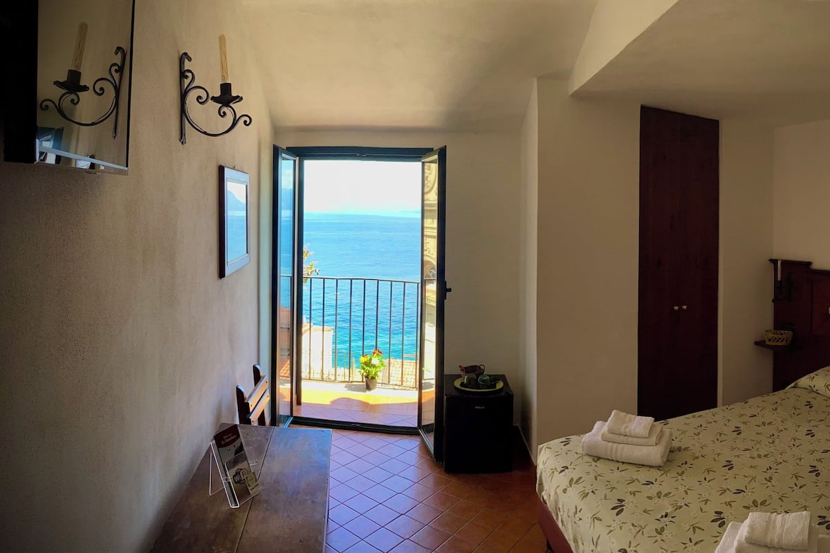 A bedroom with a bed, a door, a small table with a plant, and a balcony with a view of the sea.