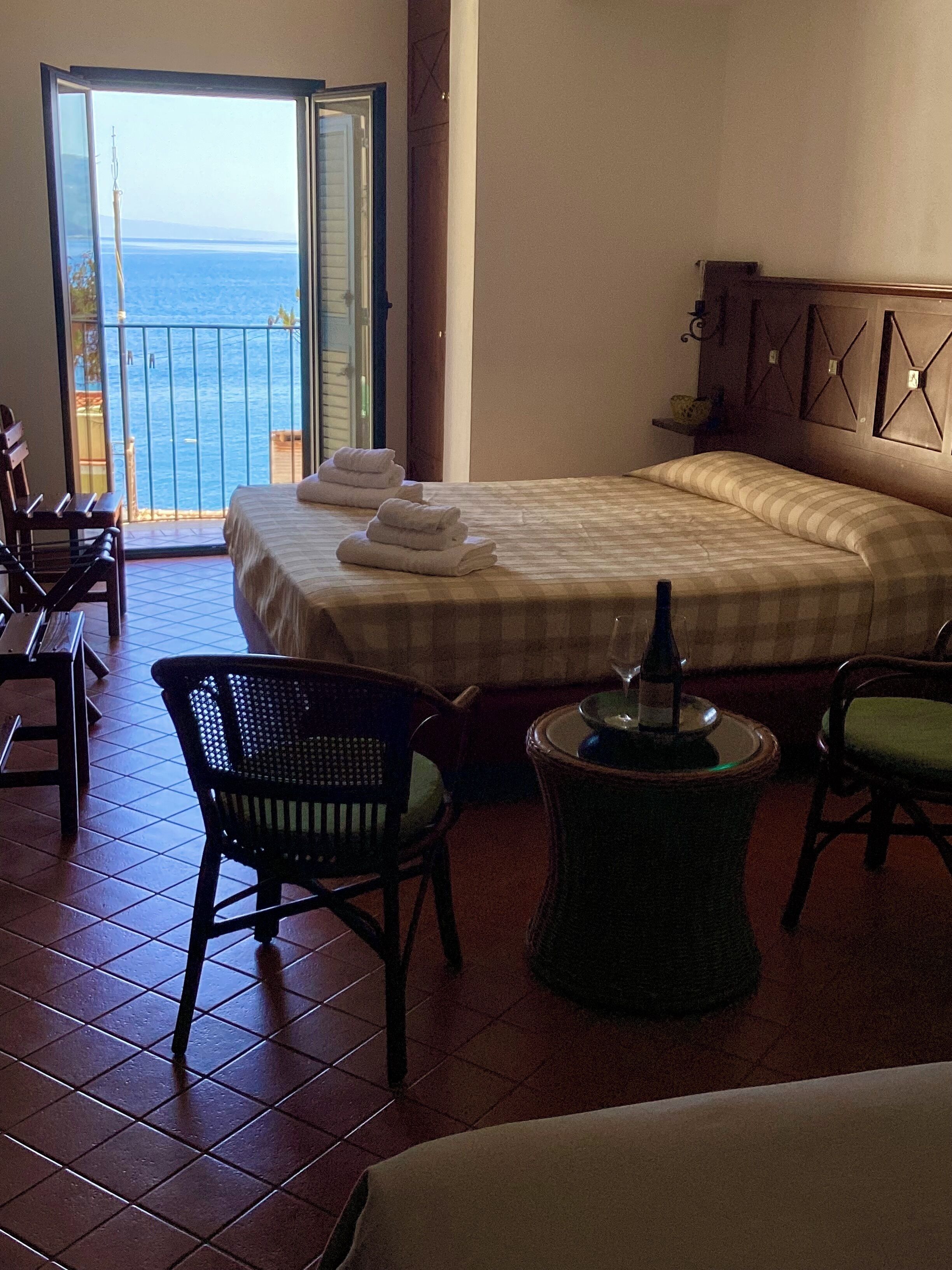 A hotel room with a bed, two chairs, a small table, and a view of the sea through an open door.