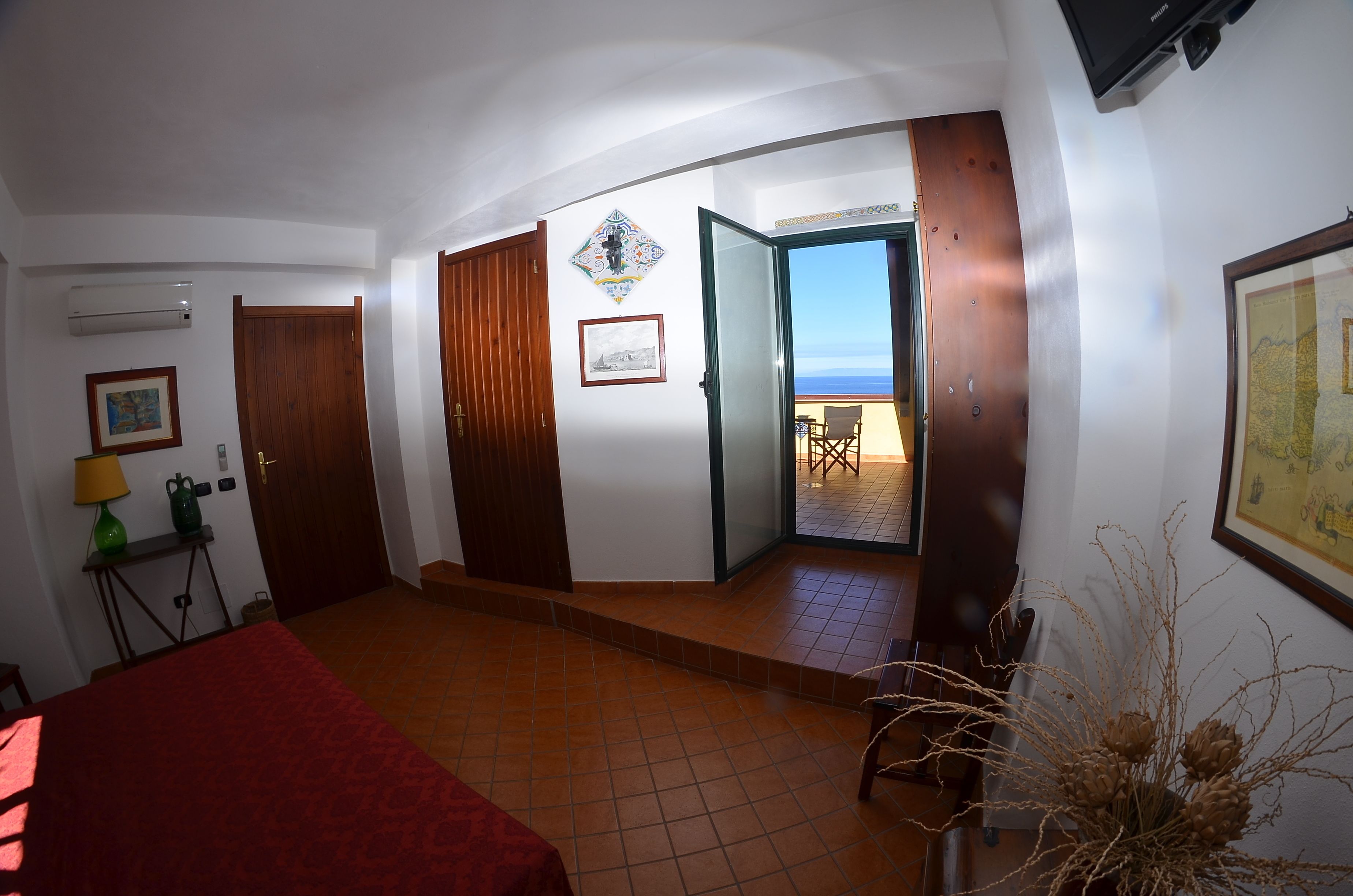 Una stanza con un letto, una scrivania, una sedia, un tavolo, una pianta e una porta che dà su un balcone con vista sul mare.