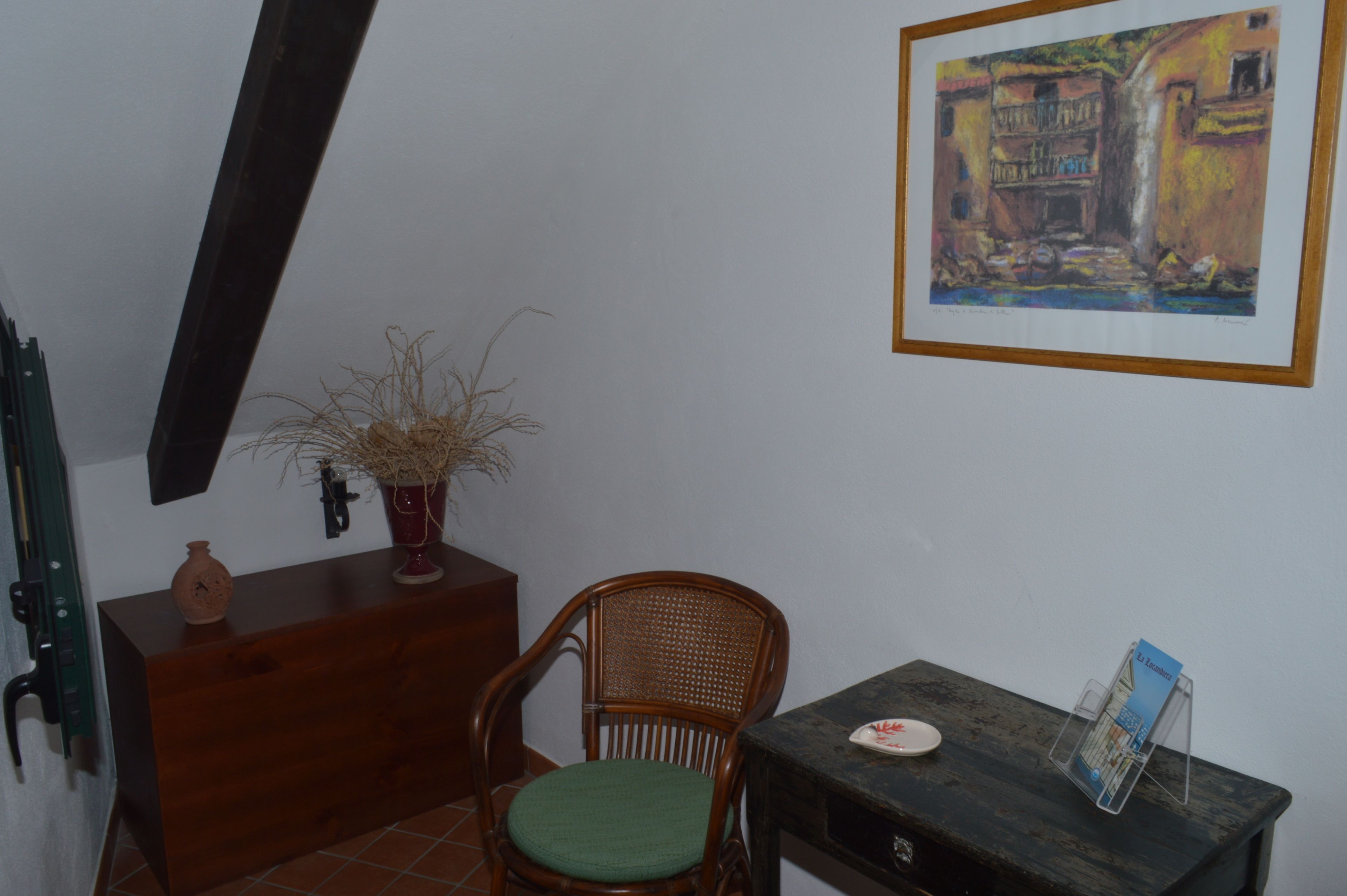 A room with a wooden chair, a small table with a brochure, a vase with dried plants, and a framed painting on the wall.