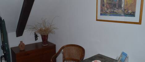 A room with a wooden chair, a small table with a brochure, a vase with dried plants, and a framed painting on the wall.