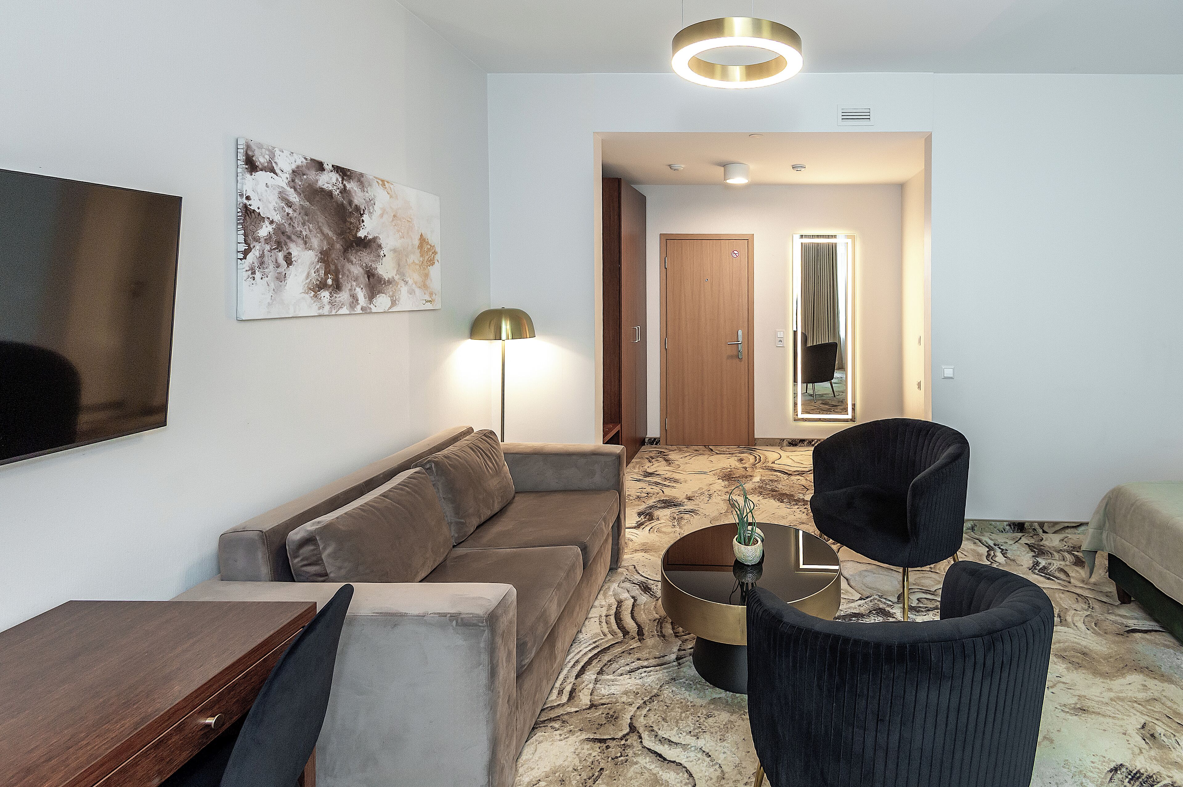 A modern hotel room with a sofa, a dining table, a black armchair, a floor lamp, and a large mirror.