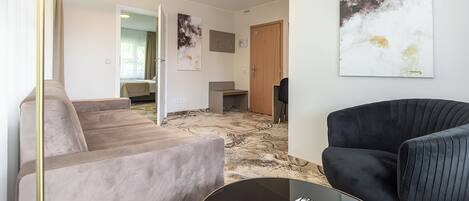 Una habitación de hotel moderna con un sofá, un sillón negro, una mesa de centro de vidrio y un libro, y arte abstracto en la pared.