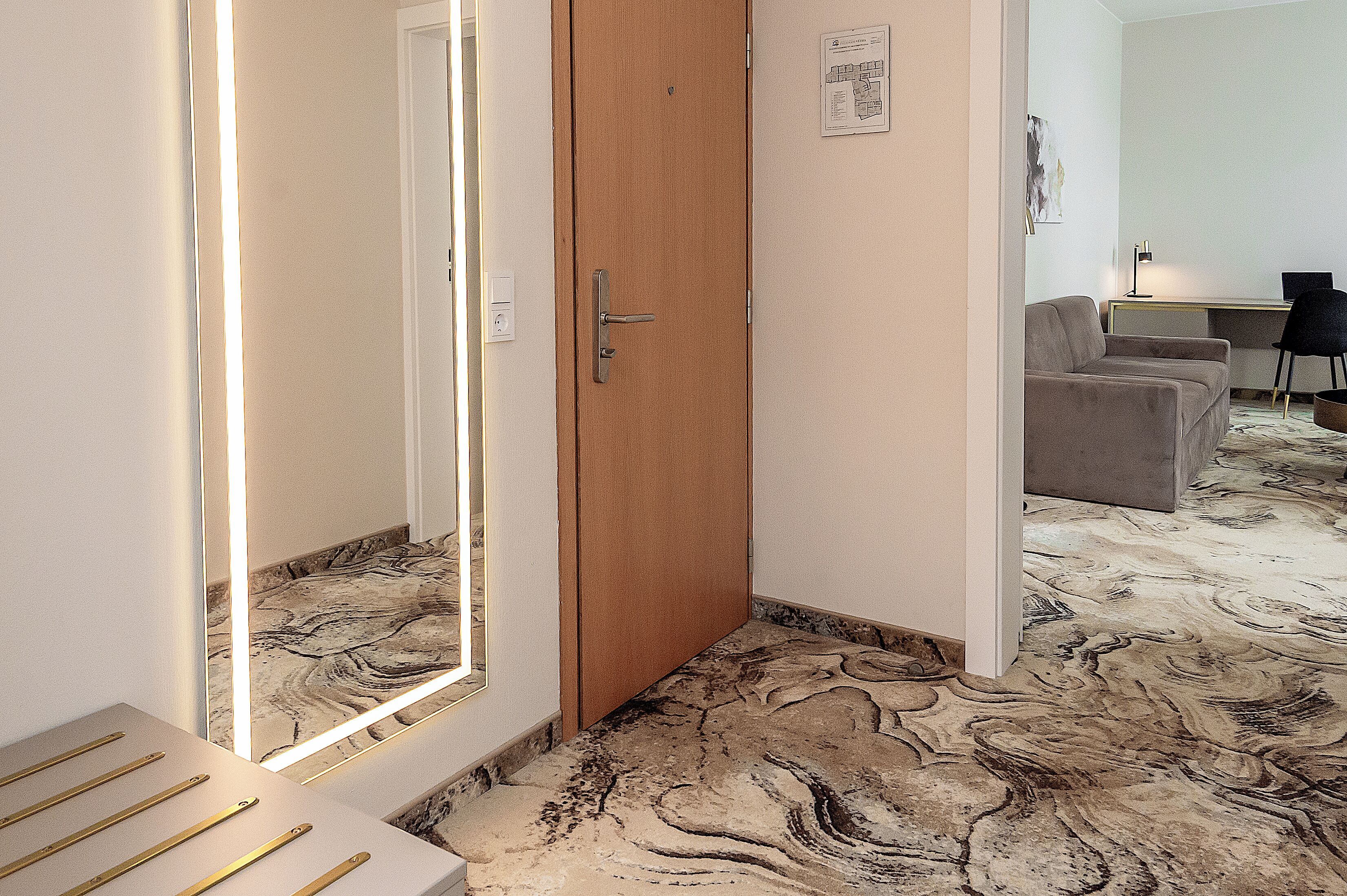 A hotel room with a wooden door, a desk with a chair, a sofa, and a patterned carpet.