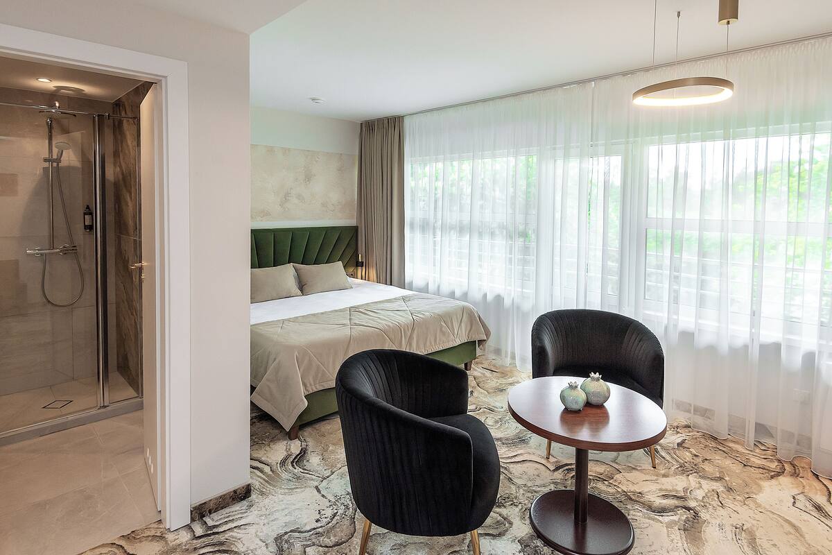 A hotel room with a shower, bed, two chairs, a table, and a view of greenery through sheer curtains.
