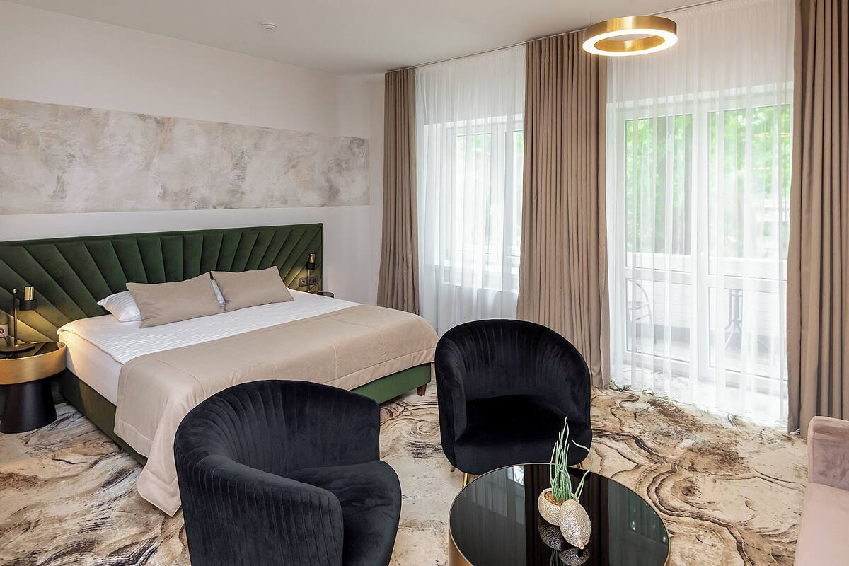 A hotel room with a large bed, two black armchairs, a round coffee table, and a view of greenery through large windows.