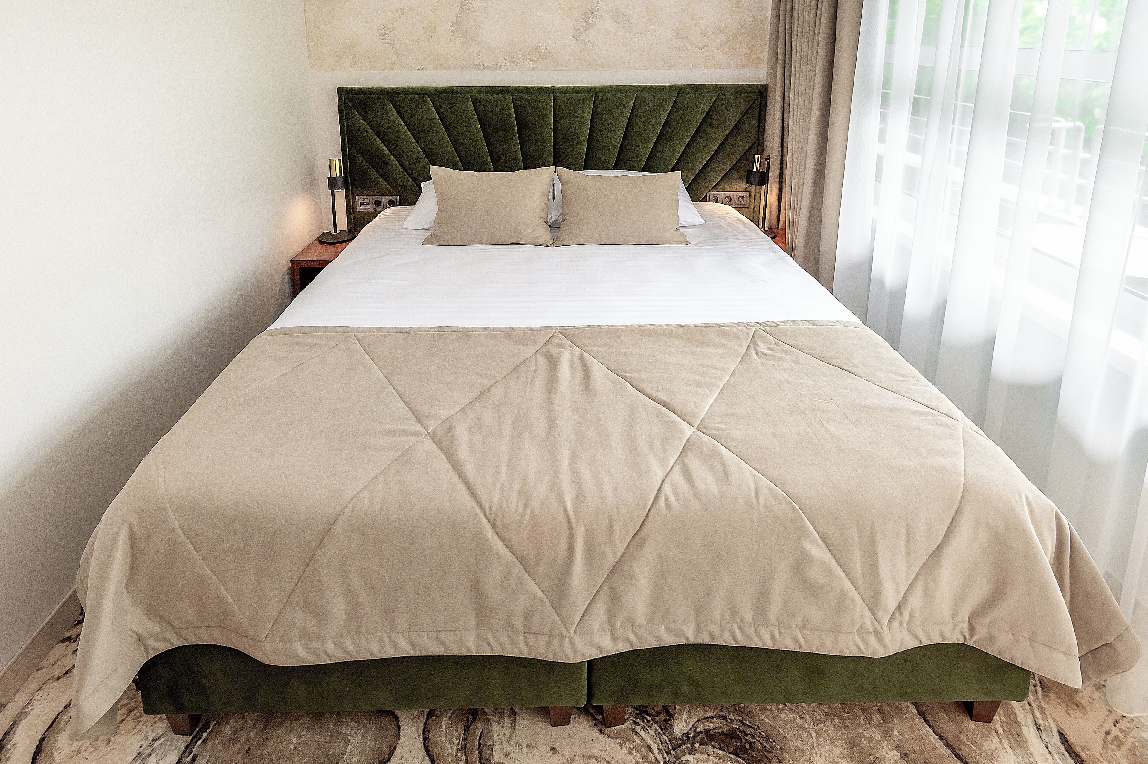 A neatly made bed with a green upholstered headboard, a beige quilted bedspread, and a wooden nightstand with a lamp.